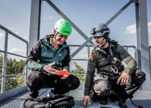 Two men in helmets on the platform of a high tower, exchanging a miniature vital signs monitor, one wearing a green helmet.