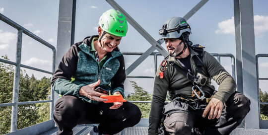 Two men in helmets on the platform of a high tower, exchanging a miniature vital signs monitor, one wearing a green helmet.